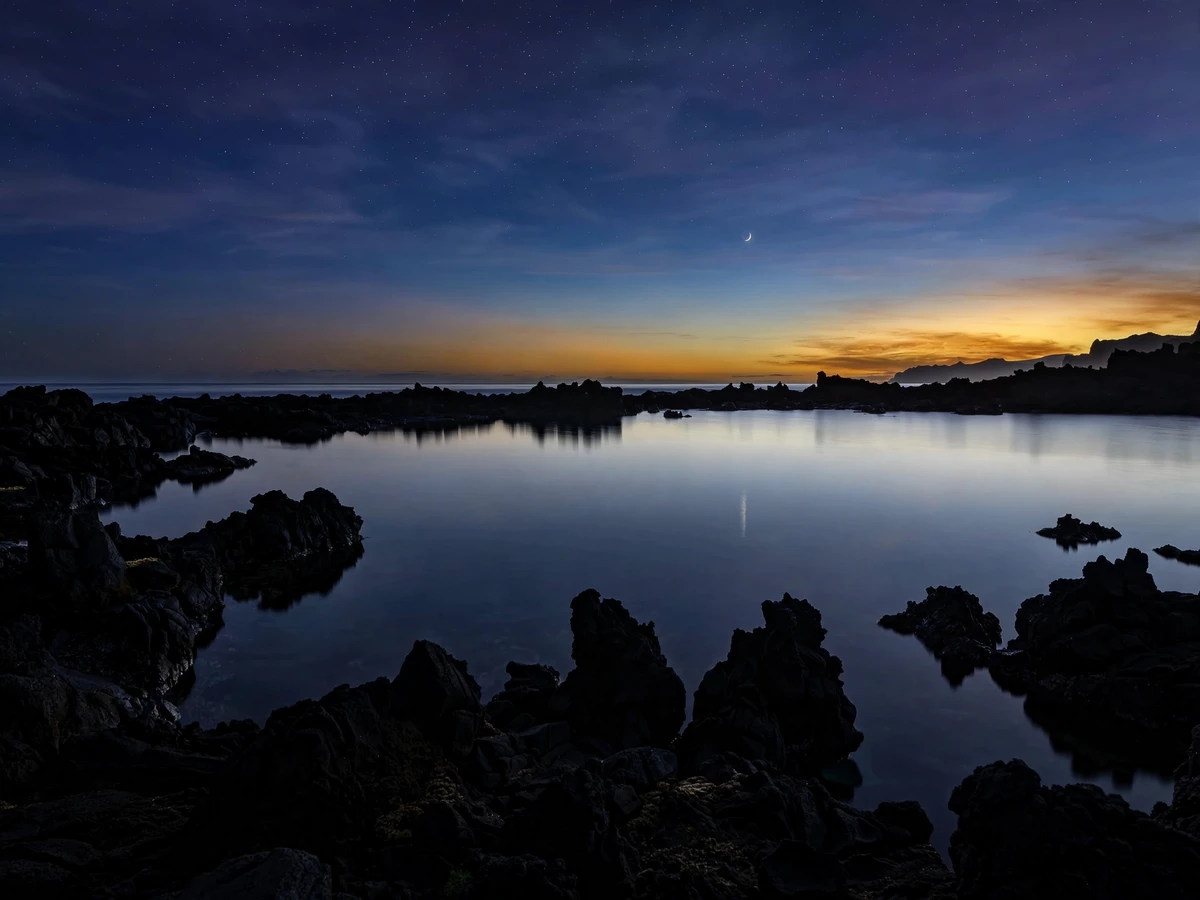Rocky Hawaiian shoreline at blue hour, calm ocean reflecting the first evening stars, horizon line sharp